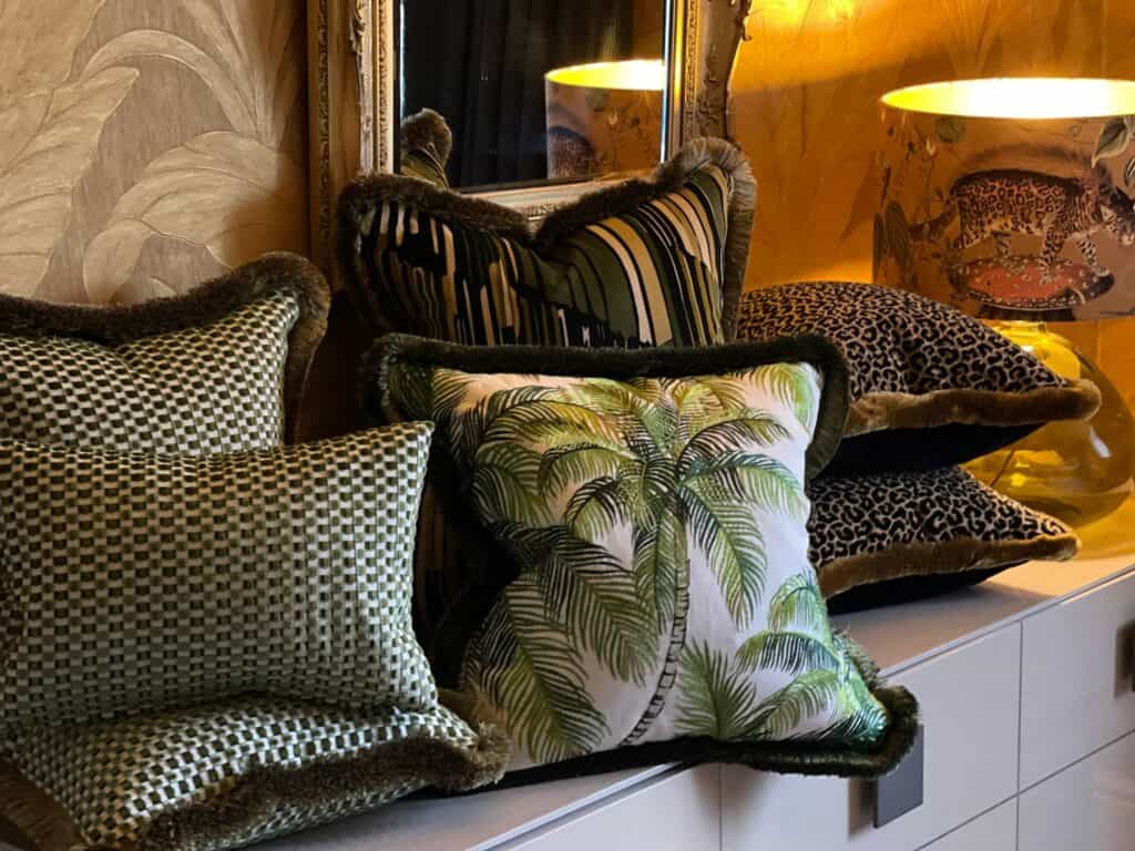 3 textures cushions with trim, 1 palm leaf cushion, 2 leopard cushions, 1 abstract cushion in front of gold trimmed mirror and Lamp